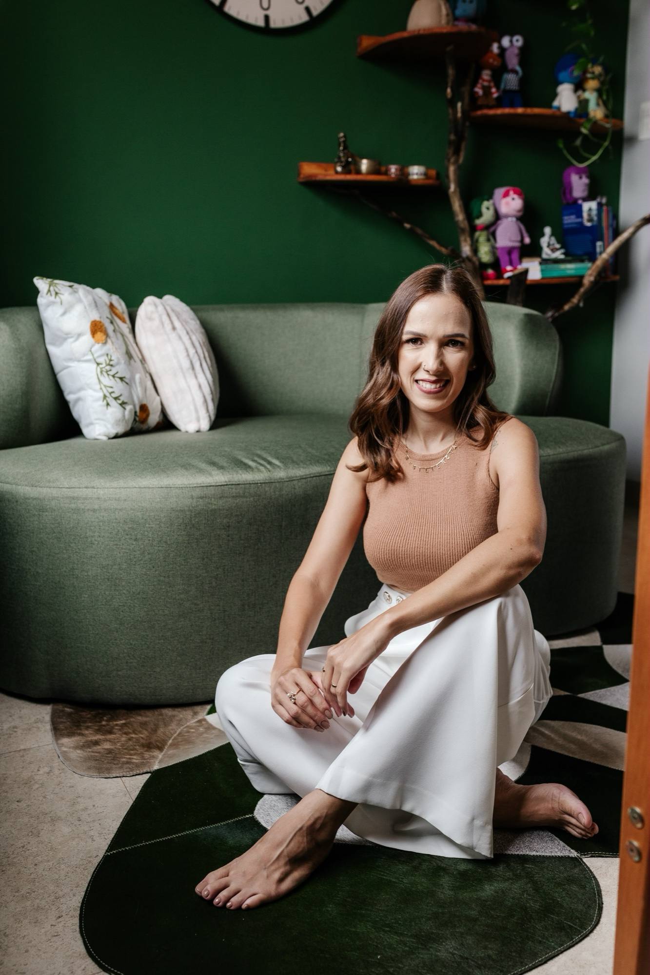 Daniela Maria sentada no chão da sala de atendimento, diante de um sofá verde-sálvia e de uma parede verde-escura com estantes.