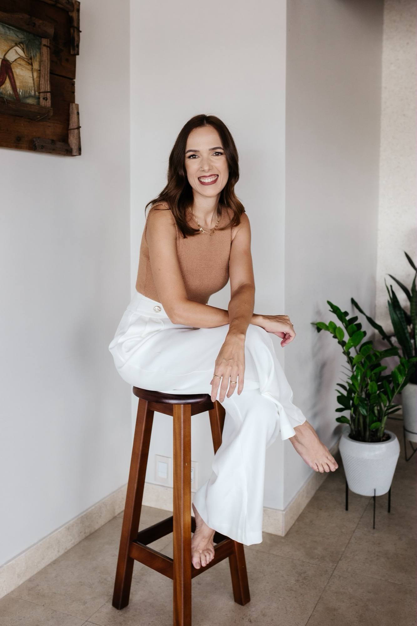 Daniela Maria sentada numa banqueta de madeira, de corpo inteiro, com blusa caramelo e calça branca, ao lado de uma planta.