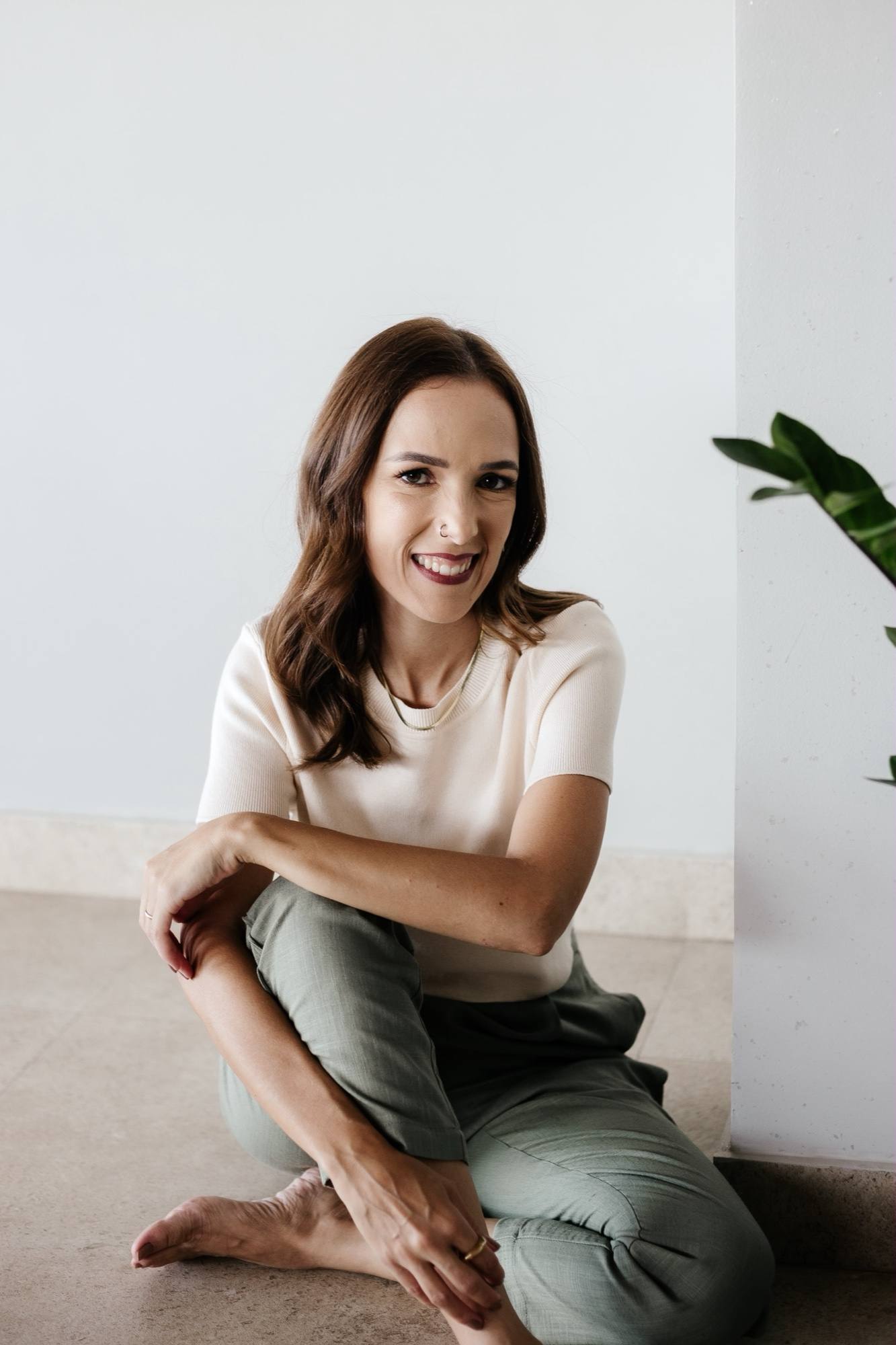 Daniela Maria sentada no chão, de blusa creme e calça verde, sorrindo, ao lado de uma planta.