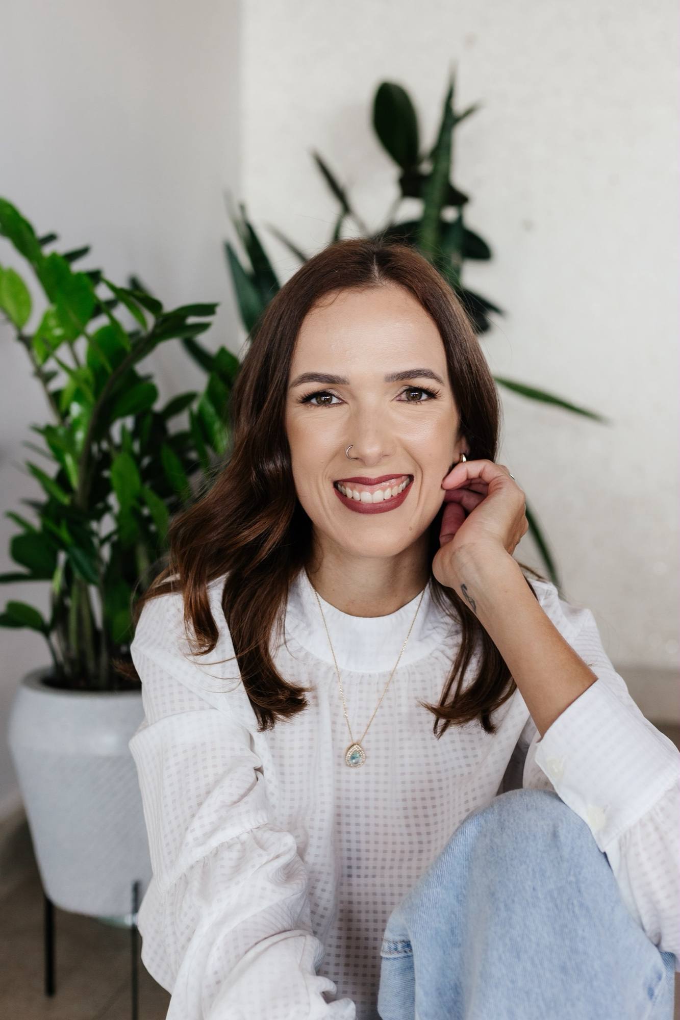 Retrato de Daniela Maria sorrindo, com a mão no rosto, entre plantas.