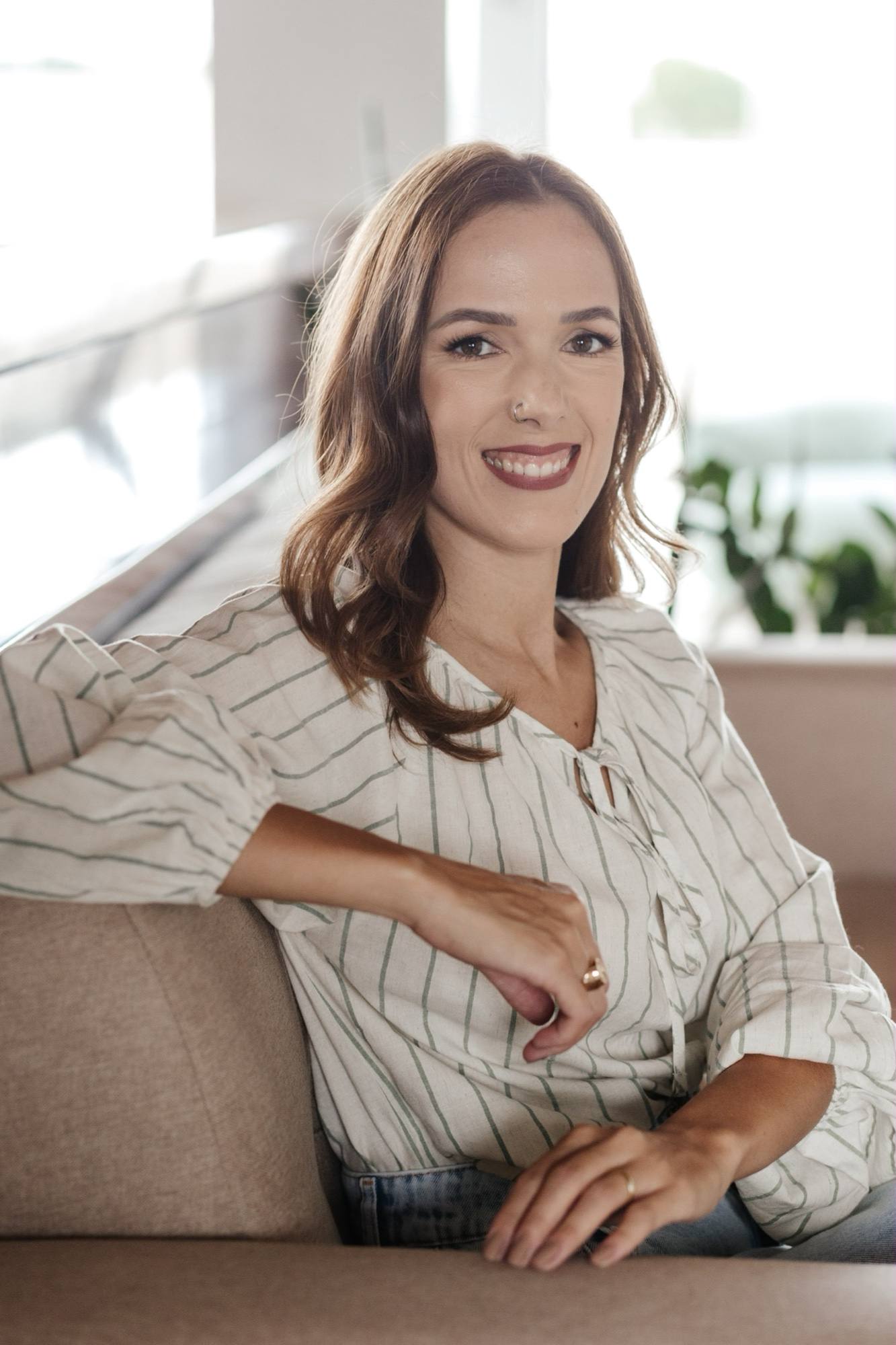 Daniela Maria sorrindo, sentada num sofá claro, de blusa listrada, com uma janela iluminada ao fundo.